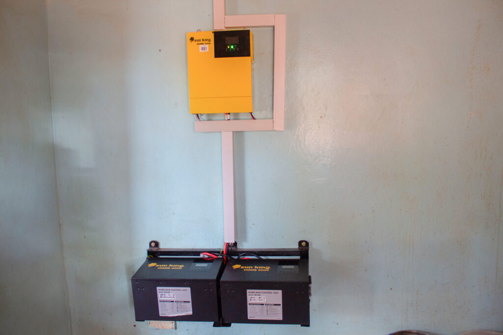 A mounted solar power system featuring a yellow Sun King inverter and two black battery control units fixed to a light blue wall, neatly connected by concealed wiring.
