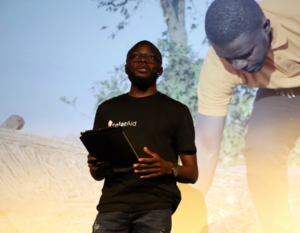 Thomas Nyangulu Jr. wearing a SolarAid T-shirt speaks while holding a tablet, standing in front of a large projected image showing a man working outdoors.