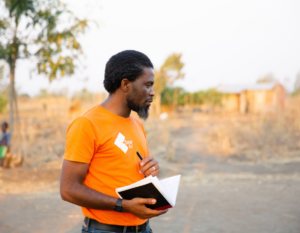 Brave Mhonie holds a notebook while working outdoors in a rural community.