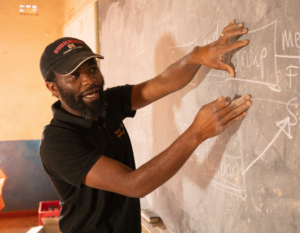 Brave Mhonie demonstrates a lesson at a chalkboard.