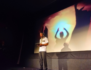 Brave Mhonie wearing a SolarAid shirt speaks on stage while holding a tablet, with a large projected image of a person raising their arms behind him.