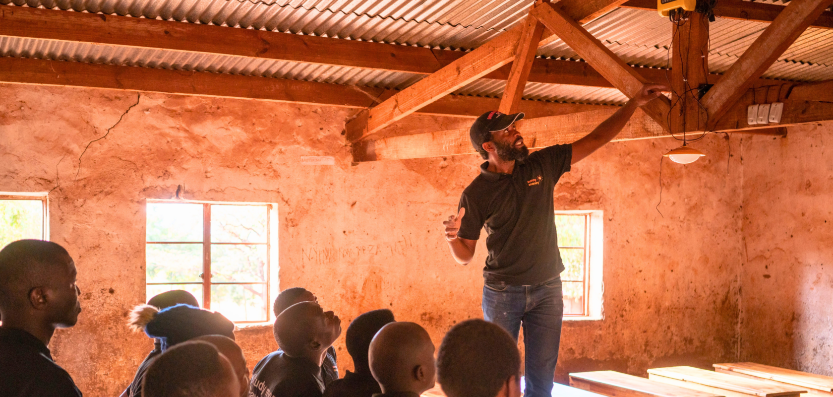 Brave Mhonie standing on a desk in a classroom, surrounded by people looking up at him, as he points towards a solar light hanging from the ceiling.