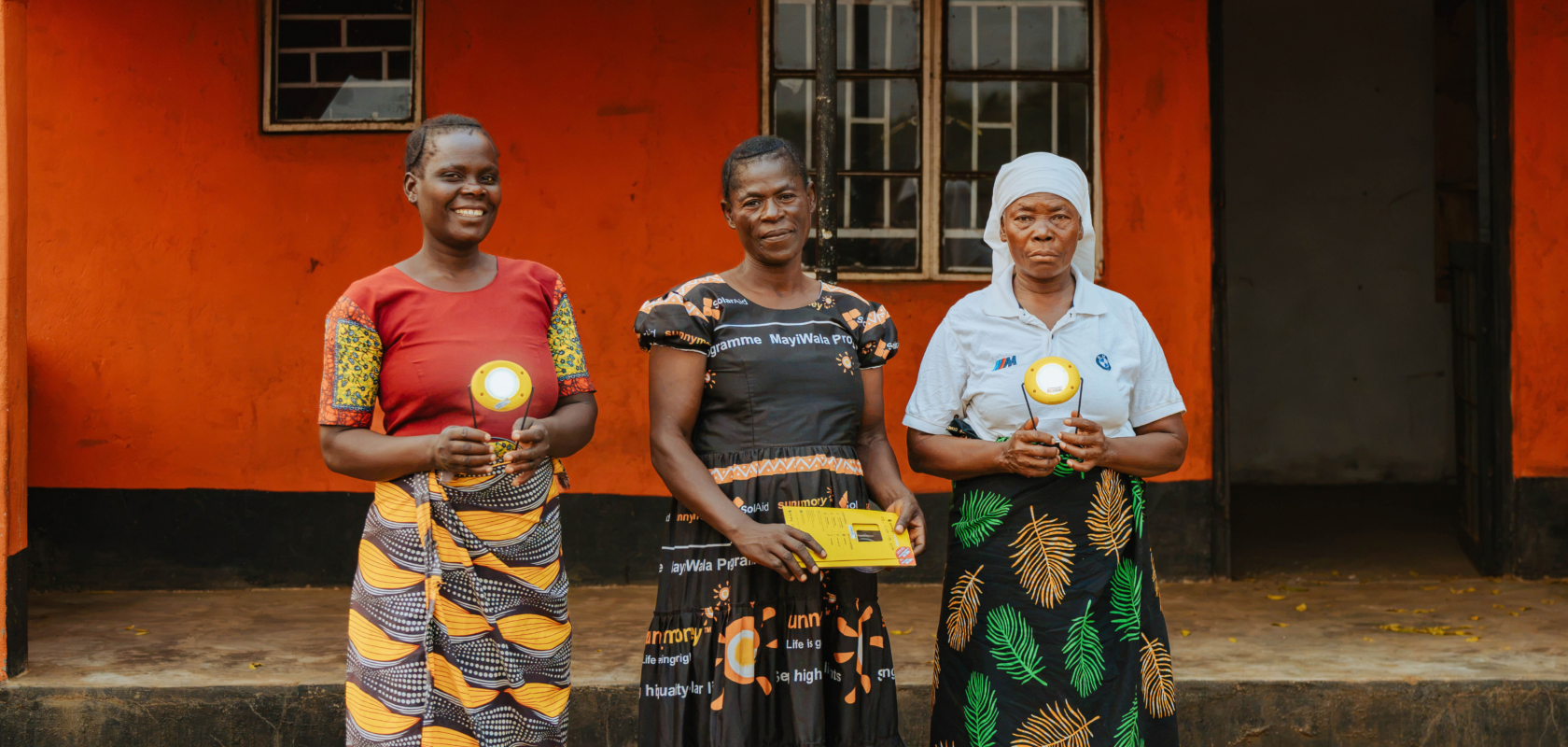Three women stand outside the orange-painted Aunt Nancy Energy Access Hub, holding solar lights.